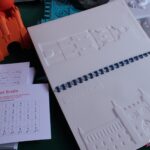 Ilustracja dotykowa Zamku Lubelskiego w książce pt. "Lublin. Przewodnik z ilustracjami dotykowymi. Ilustracje. Tom 2 z 2"; obok leżą wydruki z Alfabetem Brajla