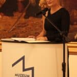 Pani Katarzyna Mieczkowska, Dyrektor Muzeum Narodowego w Lublinie, podczas wernisażu wystawy „Magia Starożytnego Egiptu”, stojąca za kremowym pulpitem z granatowym logo muzeum, za Jej plecami widoczny niewielki fragment obrazu Jana Matejki „Unia Lubelska”