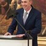 Pan Maciej Drewniak, pomysłodawca i kurator wystawy „Magia Starożytnego Egiptu”, uśmiechnięty mężczyzna w granatowym garniturze, stojący za kremowym pulpitem, za Jego plecami widoczny niewielki fragment obrazu Jana Matejki „Unia Lubelska”