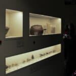 Po prawej stronie stojąca w cieniu kobieta, robiąca telefonem komórkowym zdjęcie eksponatów wystawowych