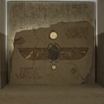 Podświetlony kamień z malowidłami i hieroglifami, na środku symbol skarabeusza uskrzydlonego