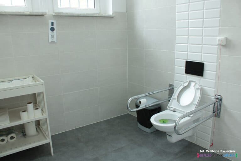 Dostępna dla wszystkich łazienka, przystosowana do potrzeb osób z niepełnosprawnościami, wyposażona w uchwyty zamontowane w pobliżu sedesu oraz system przyzywowy