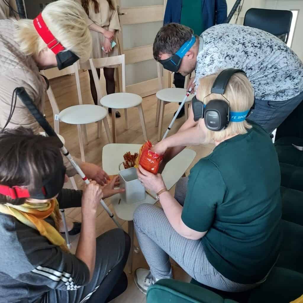 Zajęcia symulujące niepełnosprawność sprzężoną: Osoby w słuchawkach wyciszających oraz okularach zaciemniających z laskami wykonują proste czynności codzienne