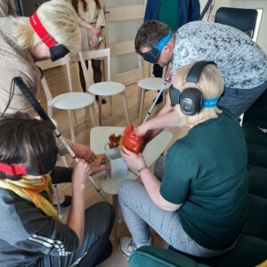 Zajęcia symulujące niepełnosprawność sprzężoną: Osoby w słuchawkach wyciszających oraz okularach zaciemniających z laskami wykonują proste czynności codzienne