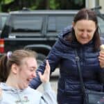 Dwie roześmiane kobiety; pierwsza siedzi na wózku, druga pochyla się nad nią, w ręku trzyma loda