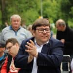 Klaszczący mężczyzna w granatowym garniturze, ma zmrużone oczy