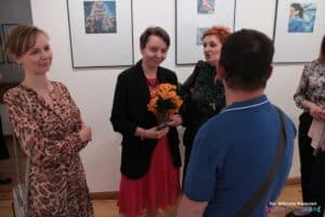 Pani Urszula Porębska trzyma pomarańczowe kwiaty, stoi wśród osób przybyłych na wernisaż, na ścianach wiszą wykonane przez Nią prace