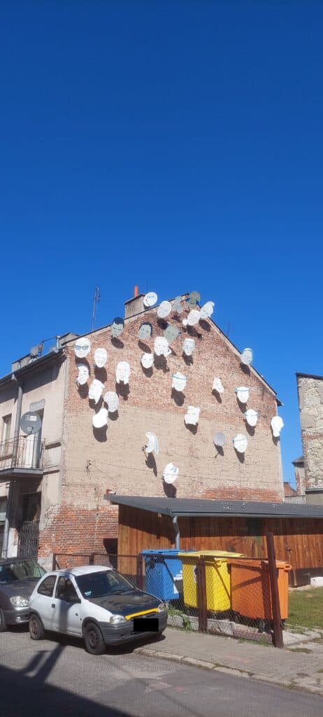 Budynek naprzeciwko lokalu Punktu Kultury. Na ścianie budynku znajdują się rzeźby twarzy w kształcie talerzy od anten satelitarnych.