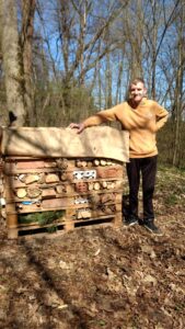 Młody uśmiechnięty mężczyzna opiera się o domek dla dzikich pszczół i innych owadów i pozuje do zdjęcia