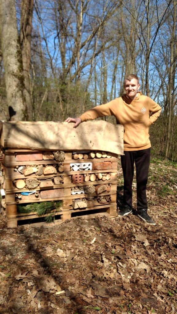 Młody uśmiechnięty mężczyzna opiera się o domek dla dzikich pszczół i innych owadów i pozuje do zdjęcia