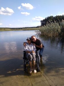 Sylwester Zams z podpiecznym podczas wakacji jednego dnia. Wraz z mężczyzną poruszającym sie na wóżku stoją na brzegu zbiornika wodnego, mają zanurzone w wodzie nogi. słoneczna letnia pogoda.