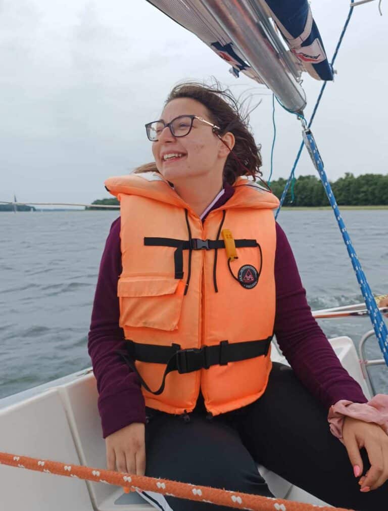 Izabela Wiater- młoda, uśmiechnięta kobieta w okularach i włosach upiętych z tyłu, ubrana w pomarańczową kamizelke ratunkową siedzi na żaglówce.