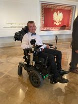 Bartłomiej Nowosadowski - mody uśiechnięty mężczyzna na wózku elektrycznym, elegancko ubrany, biała koszula, spodnie od garnituru, mucha w Wielkiej Sieni Pałacu Prezydenckiego