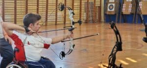 Dominik Jaszak - nastolatek z ciemnymi zaczesanymi do góry włosami. Siedzi na wózku o mierzy z łuku