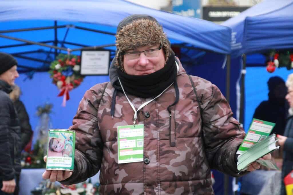 Bohater tekstu, Piotr Wróbel, ubrany w czapke, szlaik i kurtkę zimową we wzór moro.Na szyi ma identyfikator, w prawej ręce puszkę na datki, w lewej plik ulotek