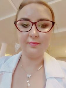 Wiola Story: młoda kobieta w dużych, brązowyh okularach, makijażu, włosy zaczesane w kok, ubrana w biału żakiet, na dekolcie naszyjnik z perełkami i motylkami.