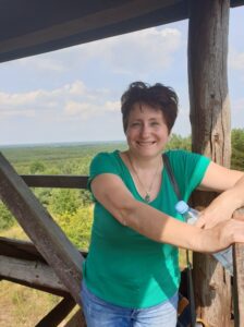 Krystyna Borusiewicz - kobieta w średnim wieku, którtkie ciemne włosy, usmiecha się do obiektywu, ubrana w zieloną koszulkę z krótkim rękawem, stoi na drewnianej wiezy widokowej oparta o barierkę. w tle słoneczne niebo i nasłonecznione lasy.