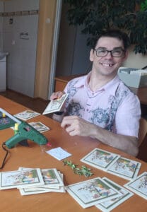 Krzysztof Warzocha- uśmiechenięty młody człowiek w okularach siedzi za biurkiem, w ręku trzyma wykonywaną przez siebie kartkę, na stole leży pistolet do klejenia