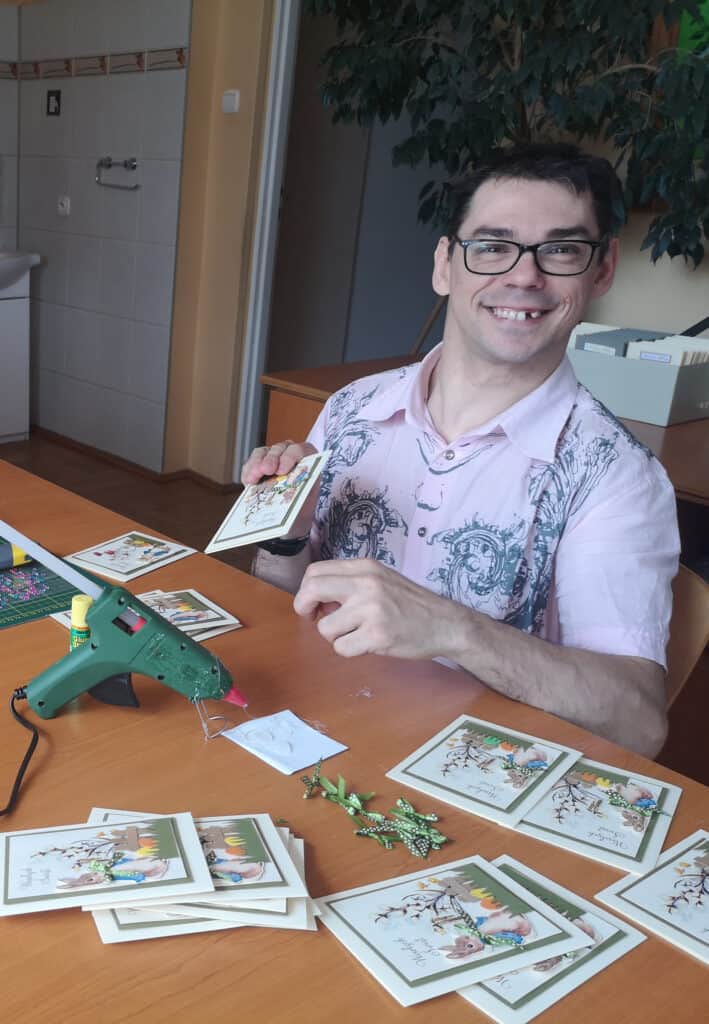 Krzysztof Warzocha- uśmiechenięty młody człowiek w okularach siedzi za biurkiem, w ręku trzyma wykonywaną przez siebie kartkę, na stole leży pistolet do klejenia