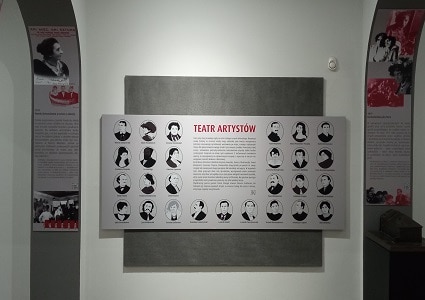 Zdjęcie ściany z tablicą, na której w centrum znajduje się tekst zatytułowany: „Teatr Artystów”. A po bokach i pod tekstem znajdują się portrety dwudziestu siedmiu osób ujęte w eliptyczne ramki. Po bokach zdjęcie podłużne pionowe tablice z grafikami i tekstami w neutralnych kolorach z dodatkiem czerwieni na szarym tle. Po prawej u dołu makieta budynku Teatru.