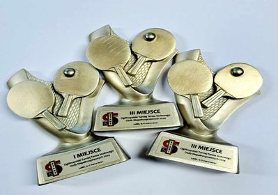 Trzy trofea w kształcie 2 rakietek i piłeczki. Każde z nich umieszczone jest na prostokątnej podstawie, na której znajduje się tabliczka z oznaczeniem miejsc: I MIEJSCE, II MIEJSCE, i III MIEJSCE.