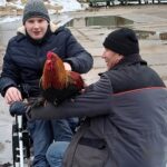 Dwóch mężczyzn, z których jeden siedzi na wózku inwalidzkim, a drugi kuca obok. Osoba kucająca trzyma koguta, który siedzi na jej ramieniu. W tle widoczny śnieg i kałuże.