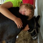 Chłopiec pochylony przytula się policzkiem do grzbietu osła w stajni. W tle widać częściowo drugą osobę w czerwonym ubraniu.