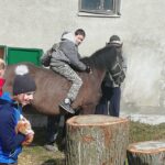 Chłopiec siedzący na koniu, którego prowadzi mężczyzna, trzymający go za uzdę. W lewym dolnym rogu zdjęcia widać dziewczynkę trzymającą rudego kota. Na pierwszym planie znajdują się pnie drzew ucięte na różnej wysokości.