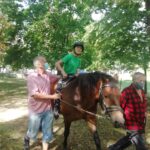 Młody jeździec, z kaskiem na głowie, siedzi na koniu pod opieką swojego instruktora w masce, który trzyma go za plecy i nogę. Drugi mężczyzna, również w masce, prowadzi konia