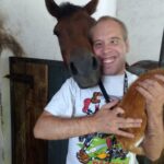 Mężczyzna uśmiecha się pozując do zdjęcia, trzymając w rękach rudego czworonoga. Z tylnej prawej strony przytula się do jego policzka koń stojący w boksie