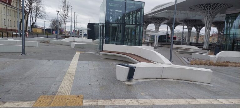 Przestrzeń miedzy "nowym" Dworcem PKS a "starym" Dworcem PKP. Na centralnym planie jasnoszare ławki. Za nimi znajduje się winda na parking podziemny.