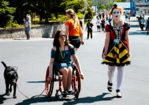 Na pierwszym planie kobieta na wózku inwalidzkim, trzymająca smycz czarnego psa. Obok niej stoi inna kobieta w makijażu jokera oraz w kolorowej, wzorzystej sukience. Obie mają identyfikatory zawieszone na szyi. W tle inni ludzie idący.