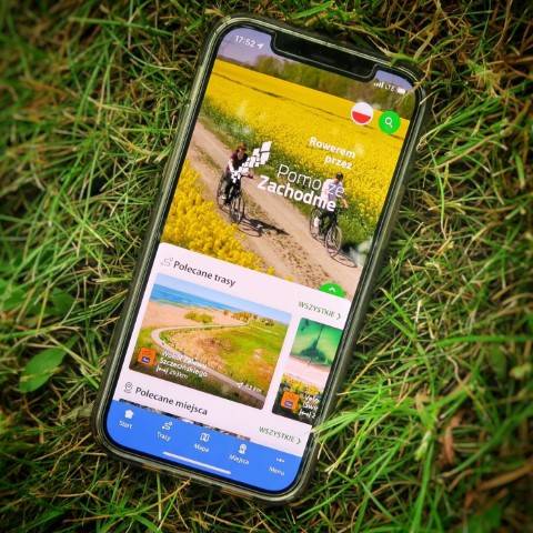 Na trawie leży telefon. Smartfon wyświetla aplikację Rowerem przez Pomorze Zachodnie z sekcjami zatytułowanymi "Polecane trasy" i "Polecane miejsca", zawierającymi miniatury krajobrazów.