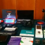 Różne urządzenia i materiały informacyjne związane z technologią wspomagającą dla osób z niepełnosprawnościami, w tym klawiatury o dużych klawiszach, specjalistyczne myszy komputerowe, tablety i inne narzędzia dostosowane do potrzeb osób z różnymi rodzajami niepełnosprawności. Na stole leżą również broszury i ulotki informacyjne dotyczące technologii wspomagającej.