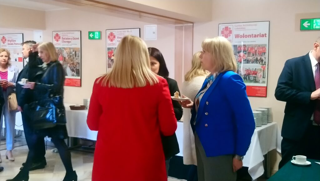 Trzy kobiety stojące w grupie - jedna z nich odwrócona jest tyłem do obiektywu, druga stoi bokiem, a twarz trzeciej kobiety częściowo wyłania się zza pierwszej. W tle zauważalni inni ludzie.