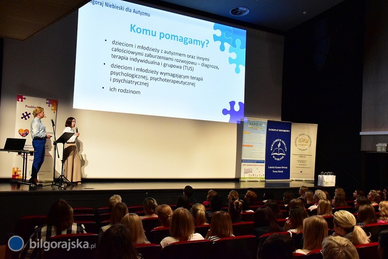 Zdjęcie prezentacji multimedialnej pt. Komu pomagamy? z obok stojącymi dwiema kobietami prezentującymi ją słuchaczom.