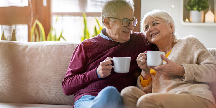 Para starszych państwa siedzi na kanapie, przytulona do siebie i trzymająca w ręku białą filiżankę