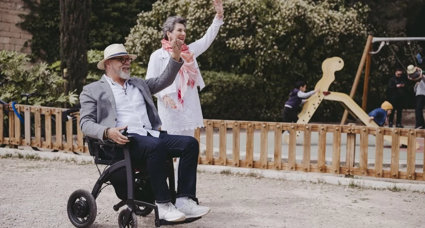 Para ludzi w średnim wieku. Mężczyzna siedzi na wózku inwalidzkim, a kobieta stoi obok niego. Oboje machają ręką. W tle widać plac zabaw ogrodzony drewnianym płotem, na którym bawią się dzieci.