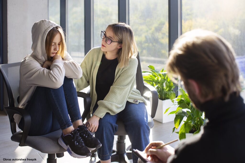 3 osoby siedzą w jasnym pomieszczeniu z dużymi oknami. Nastolatka i młoda kobieta po lewej stronie są skierowane w stronę mężczyzny po prawej stronie, który notuje w zeszycie. Nastolatka po lewej nosi bluzę z kapturem i dżinsy, a młoda kobieta w środku nosi jasnozielony sweter i dżinsy. W tle widać rośliny doniczkowe i drzewa za oknem.