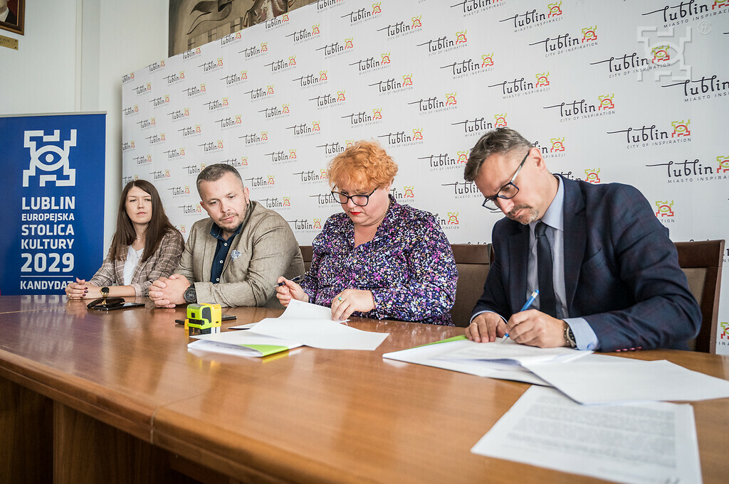 Cztery osoby siedzące przy stole podczas formalnego spotkania. Na stole leżą dokumenty, a dwie osoby podpisują je. W tle widoczne są banery z logotypem "Lublin - miasto inspiracji
