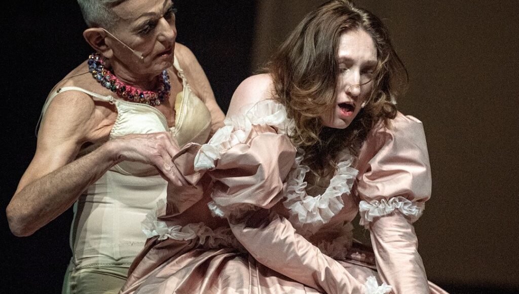 Kadr ze spektaklu: Orlando. Biografie. Dwie osoby w kostiumach, w pozach pełnych dramatyzmu. Jedna osoba pomaga drugiej włożyć epokową suknię z bufiastymi rękawami.