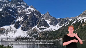 Kadr z filmu z tłumaczką języka migowego umieszczoną w dolnym prawym rogu ekranu. Widoczne są na nim strome, skaliste szczyty gór częściowo pokryte śniegiem oraz jezioro u ich podnóża. Na dole napis: Morskie Oko jest najchętniej odwiedzanym miejscem na terenie Tatrzańskiego Parku Narodowego.