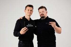 Dwóch mężczyzn, którzy obejmują się, ubrani są w czarne koszulki. Mężczyzna po lewej stronie trzyma opakowanie z napisem SuperWgistle, natomiast jego towarzysz po prawej trzyma w dłoni gwizdek.