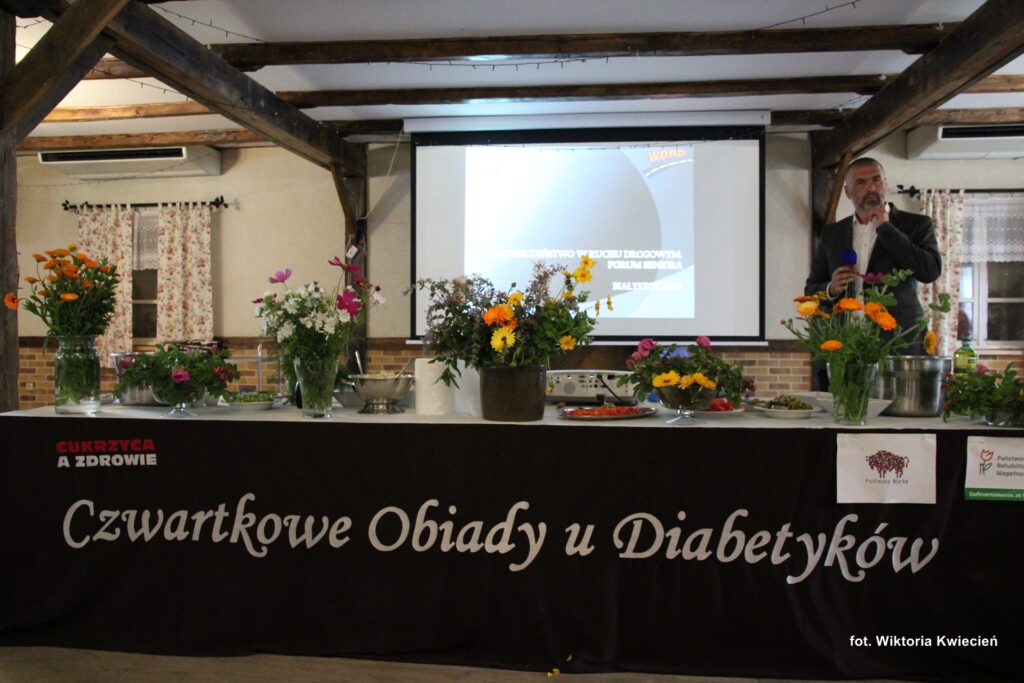 Stół nakryty brązowym obrusem z wyszytym napisem: "Czwartkowe Obiady u Diabetyków". Na stole stoją kolorowe bukiety świeżych kwiatów. Za stołem stoi Pan Paweł Dunaj, a w tle widać ekran multimedialny; fot. Wiktoria Kwiecień
