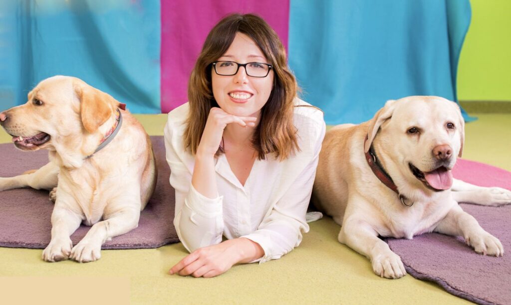 2 labradory siedzące na fioletowych matach. Psy są ustawione po obu stronach młodej kobiety w okularach.