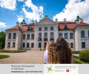 Budynek o barokowym stylu architektonicznym. Napis na dole obrazka wskazuje: Muzeum Zamoyskich w Kozłówce. Widać dwie młode dziewczyny z długimi włosami odwrócone plecami do aparatu. Na prawym dolnym rogu obrazka jest informacja, że Muzeum zostało uznane za NAJLEPSZY PRODUKT TURYSTYCZNY WOJEWÓDZTWA LUBELSKIEGO 2023.