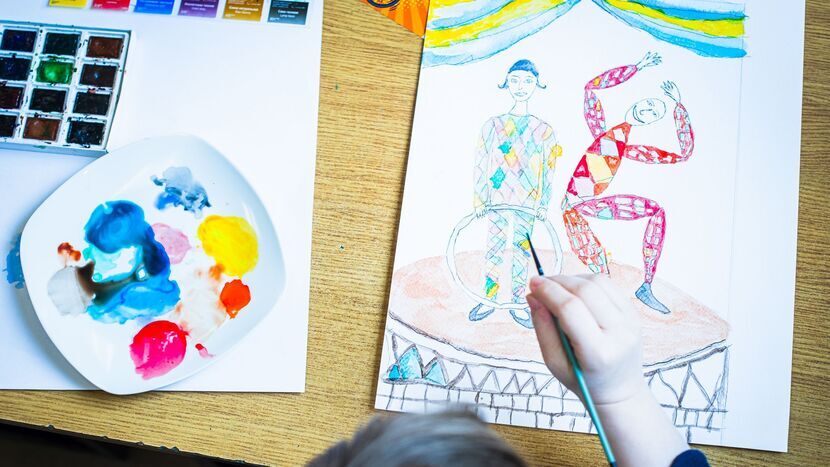 Zbliżenie na stół z artykułami do sztuki oraz rysunkiem. Po lewej stronie widoczna jest paleta z różnymi kolorami farb. Obok palety znajduje się praca artystyczna w trakcie tworzenia, przedstawiająca dwie postacie ludzkie: jedna z postaci stoi, trzymając ręce na hula hop, podczas gdy druga tańczy. Nad rysunkiem unosi się dziecięca ręka trzymająca pędzel.