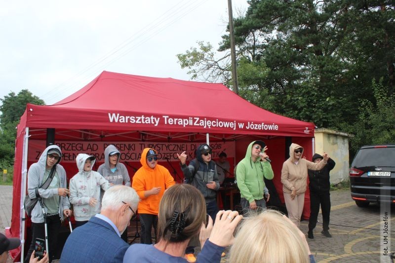 Grupa siedmiu osób w kapturach stoi pod czerwonym namiotem, na którym widnieje napis: 'Warsztaty Terapii Zajęciowej w Józefowie'. Jedna z tych osób trzyma mikrofon.