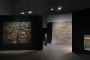 Wnętrze galerii sztuki o przyćmionym oświetleniu. Na ścianach widoczne są trzy prace artystyczne.