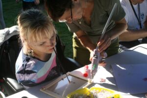 Blondynka maluje pędzlem w ustach wnętrze ramki, obok niej stoi kobieta pochylona i nakłada w ramce klej pistoletem.